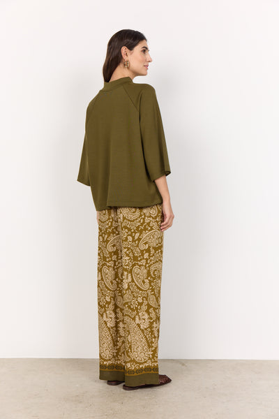 Woman wearing an olive green top and patterned pants against a white background
