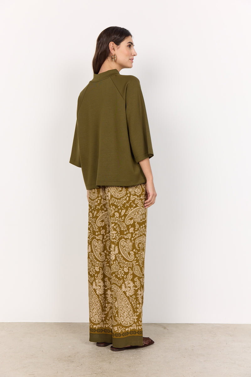 Woman wearing an olive green top and patterned pants against a white background