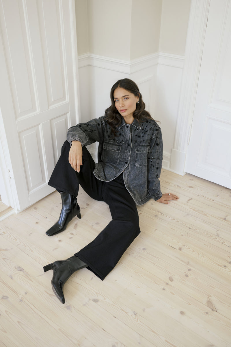 Woman sitting on a wooden floor wearing a denim jacket and black jeans.