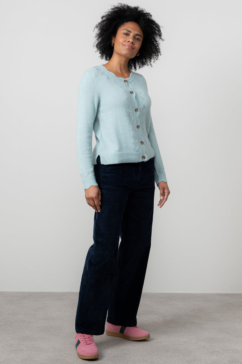 Woman wearing lily & me light blue cardigan, dark pants, and pink shoes on a plain background