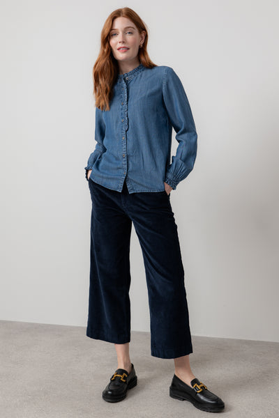 Woman wearing a blue denim shirt and dark blue pants against a plain background