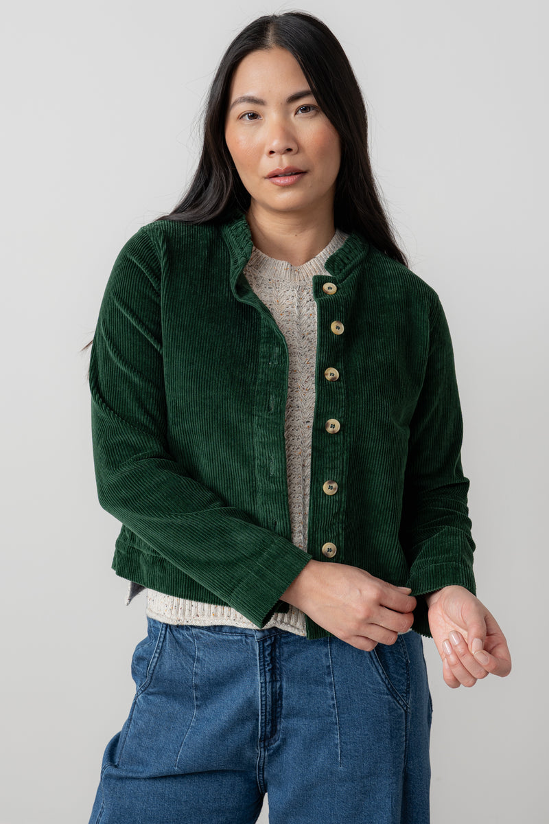 Woman wearing rene green corduroy jacket over a sweater and blue jeans on a plain background