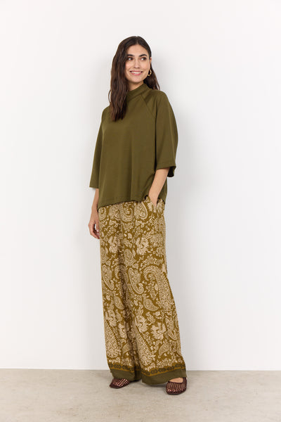 Woman wearing an olive green top and patterned pants against a white background