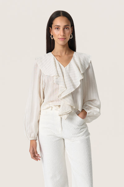 model wearing soaked in luxury roya cream blouse with fine dark stripe, soft ruffled v neck collar and long sleeves. model is wearing the blouse with cream trousers