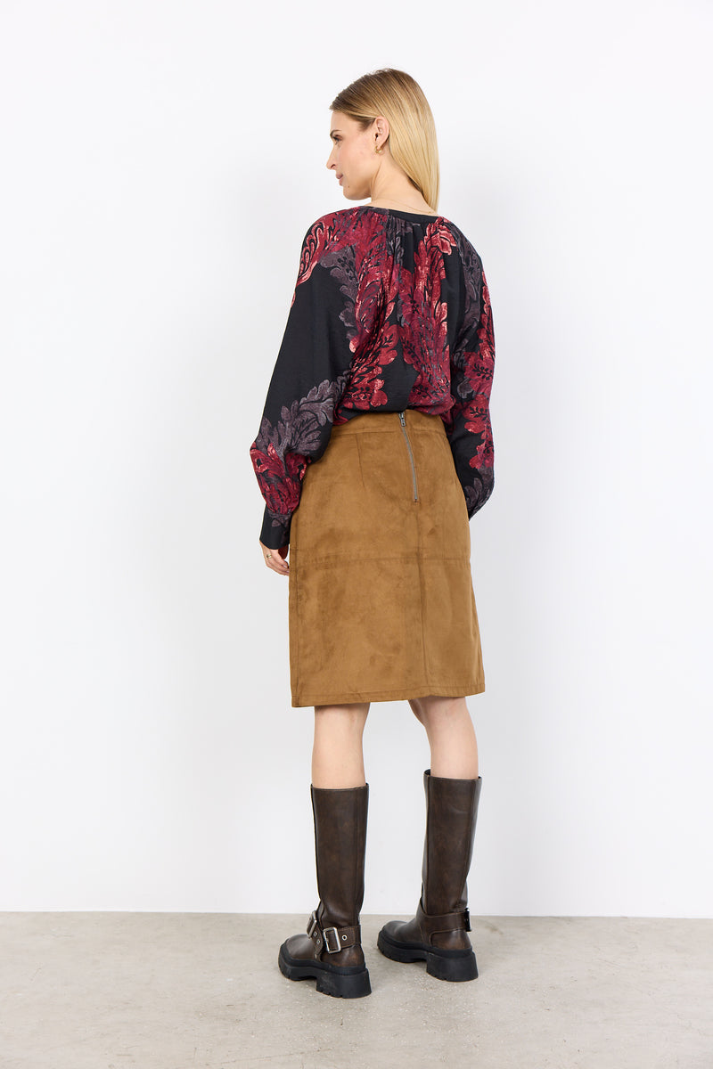 Woman wearing a patterned top, brown skirt, and black boots against a white background