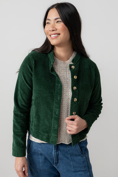 Woman wearing a green corduroy jacket over a beige sweater and blue jeans on a plain background