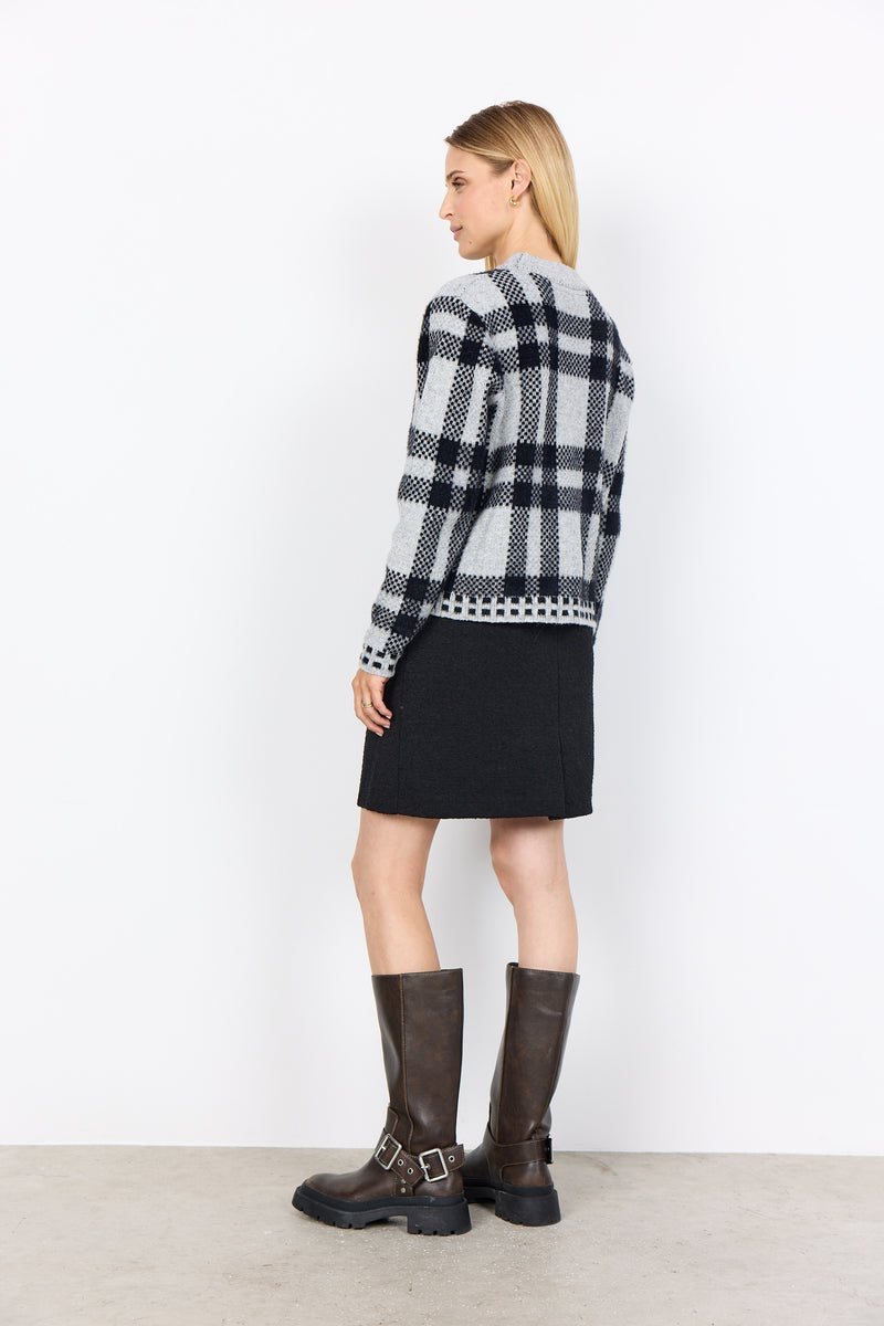 Woman wearing soyaconcept black and white checkered sweater, black skirt, and brown boots against a white background